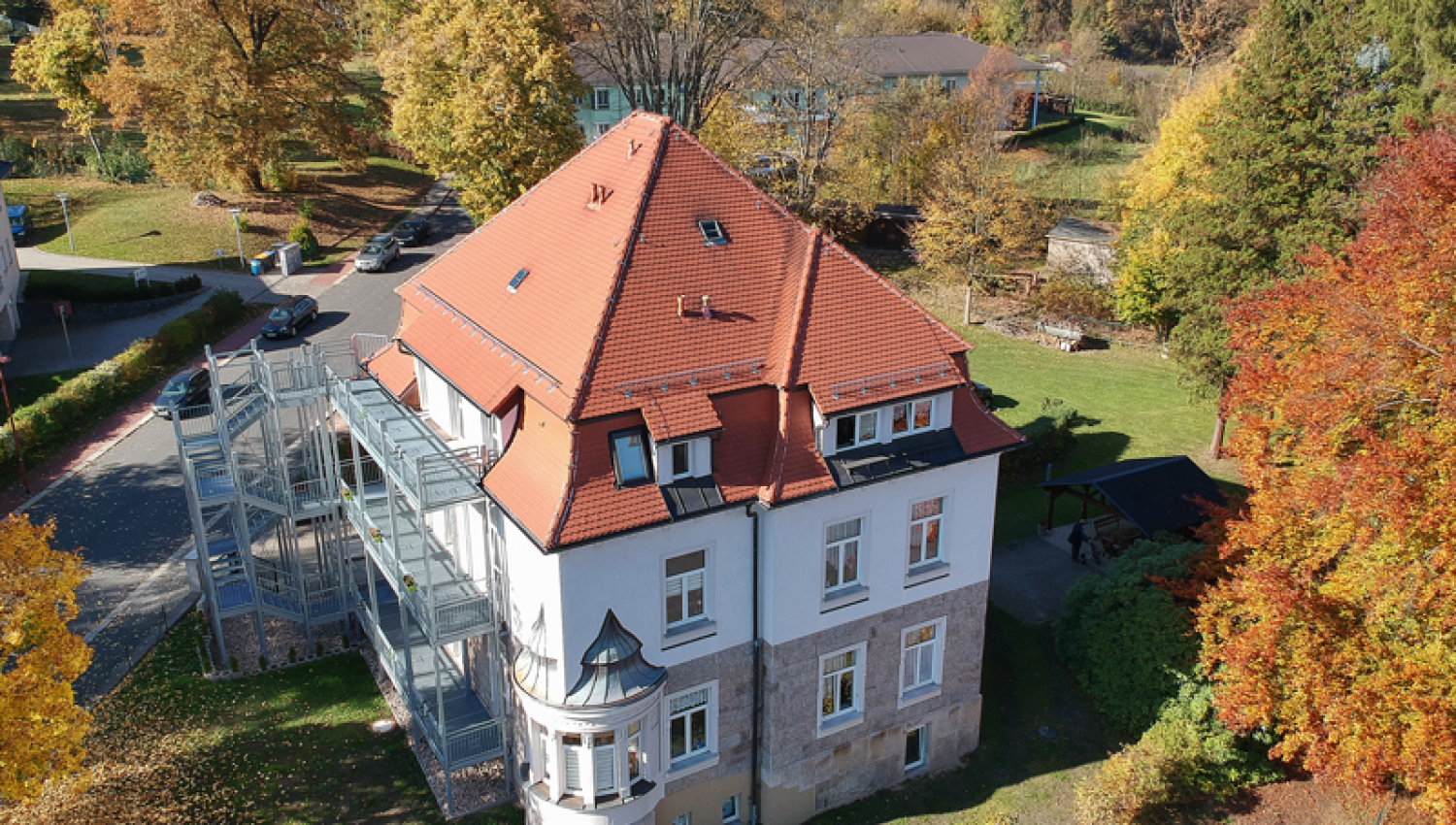 Ein großes weißes Haus mit rotem Ziegeldach und Glasanbau steht inmitten bunter Herbstbäume, im Hintergrund sind grüne Rasenflächen und weitere Gebäude zu sehen.